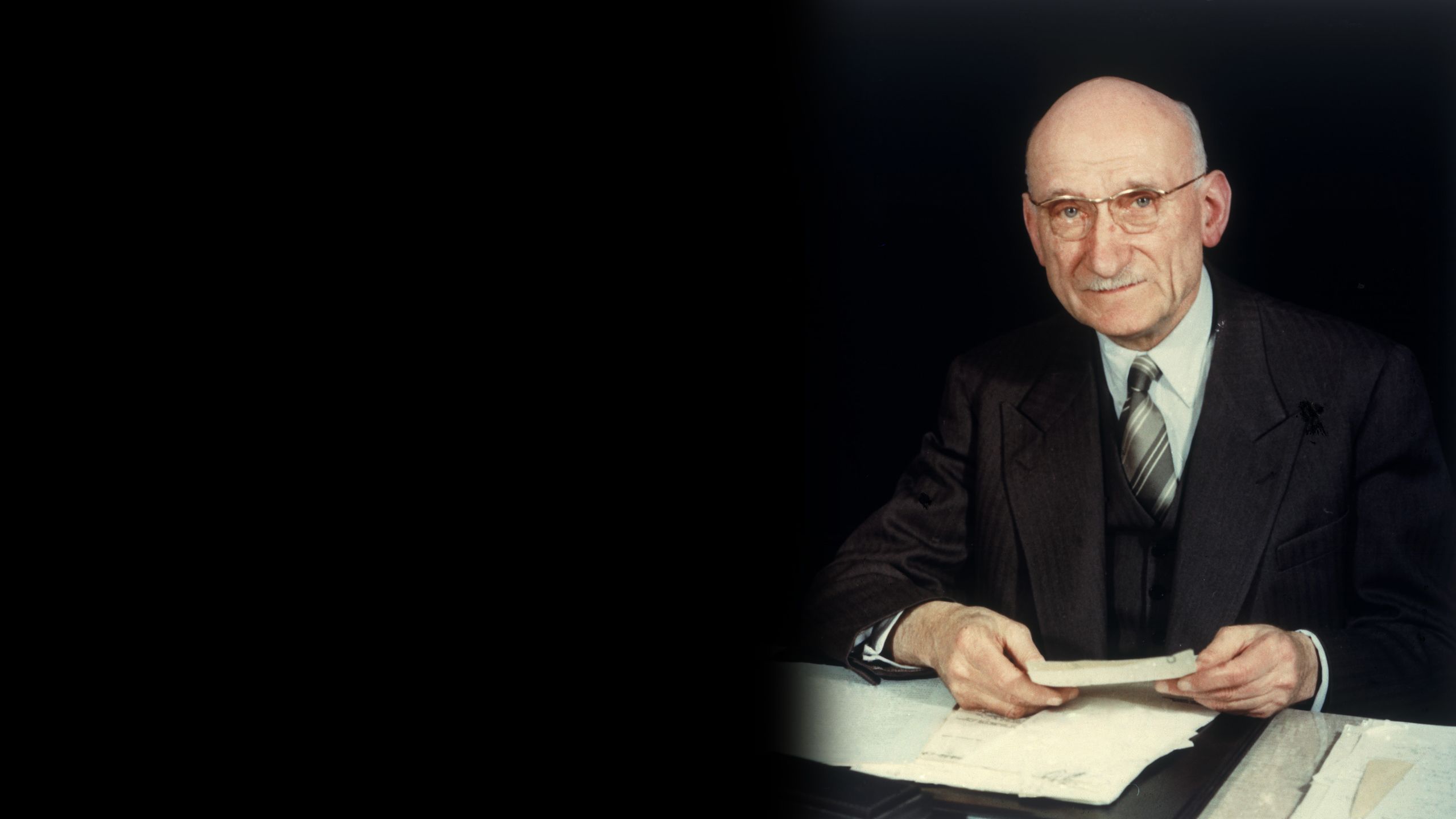 Robert Schuman sedí za stolem, dívá se do objektivu a v ruce drží dokumenty.