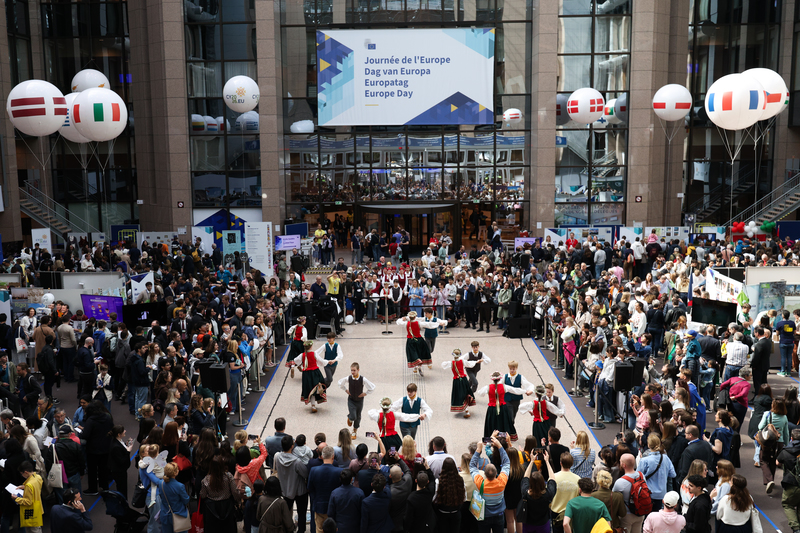 Des visiteurs sont rassemblés dans un grand hall à l'occasion de la Journée de l'Europe. Ils regardent des danseurs en costumes traditionnels se produire sur une scène centrale. L'espace est décoré de drapeaux et de ballons.