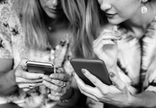 Dvě ženy sedí vedle sebe, každá drží v ruce svůj mobilní telefon a dívá se na něj.