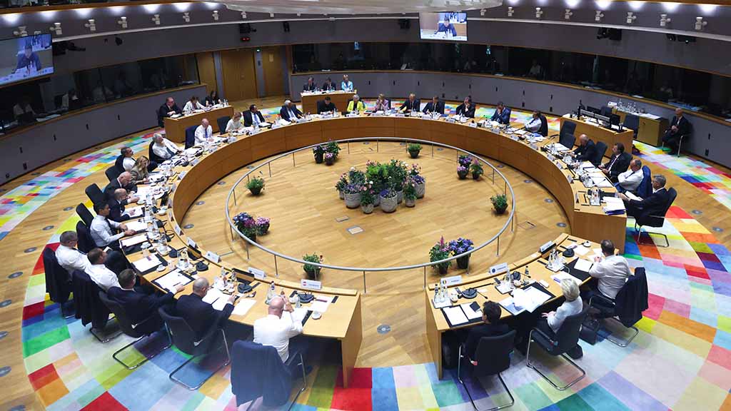 Dans une salle bien éclairée, des officiels en costume assis autour d'une grande table de conférence circulaire entourée d'une moquette aux motifs colorés, avec des plantes en pot au centre de la pièce.